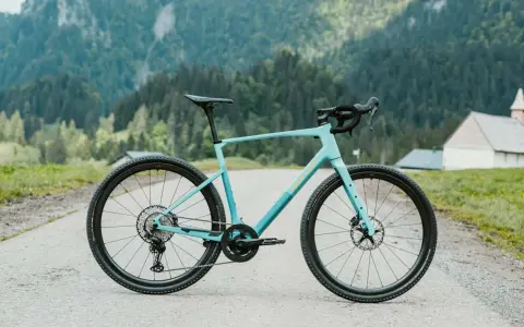 Ein hellblaues Simplon Grid Gravelbike steht auf einer asphaltierten Straße vor einer Bergkulisse mit Wäldern und einer kleinen Kirche.