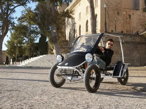 Ein Mann fährt einen Duo Moke durch eine sonnige Gasse mit historischen Gebäuden im Hintergrund.