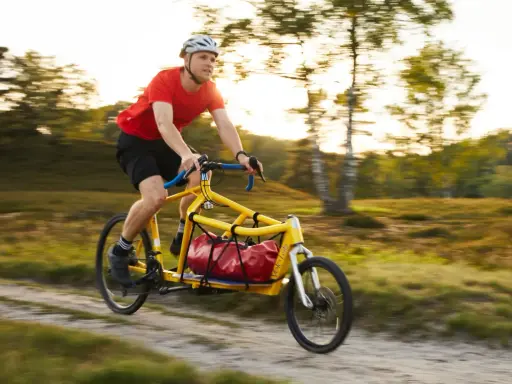 Mann fährt mit einem gelben Velo Lab Lastenrad mit roter Tasche durch eine sonnige Landschaft.