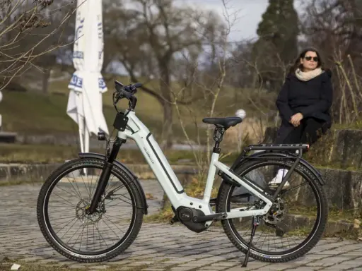 Eine Frau sitzt neben einem hellgrauen Velo de Ville, das auf einem gepflasterten Weg steht. Gute Velo de Ville Erfahrungen sind garantiert.