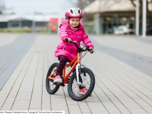 Ein fröhliches Mädchen in pinker Jacke und Helm sitzt auf einem orangefarbenen Kinderfahrrad. Die Frage "kinderfahrrad 4 jahre wieviel zoll" wird durch die Größe des Fahrrads beantwortet.