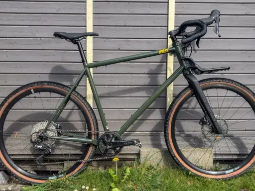 Grünes Rose Hobo Test Gravelbike mit braunen Reifen und Gepäckträger vor Holzwand.