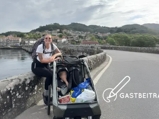 Frau mit Rucksack und Kinderwagen auf einer Brücke, bereit für eine abc tour. Im Hintergrund eine Stadt und Berge.