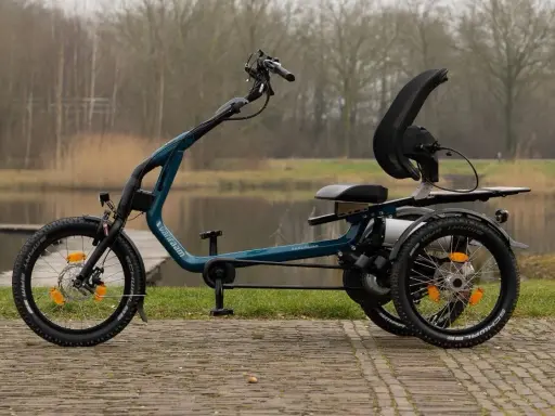 Ein tealfarbenes Easy Rider Bike mit drei Rädern steht am Wasser. Es hat einen bequemen Sitz und eine hohe Rückenlehne.