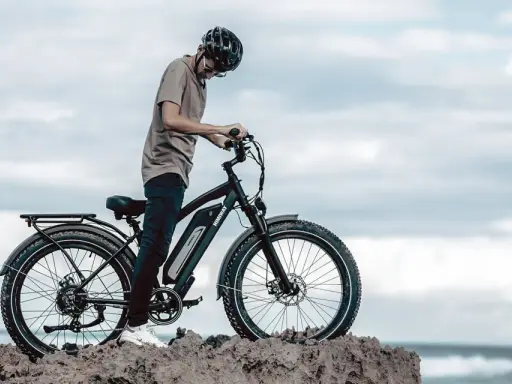 Ein Mann mit Helm steht neben einem Fatbike mit breiten Reifen am Meer. Was ist ein Fatbike? Ein robustes Fahrrad für jedes Gelände.