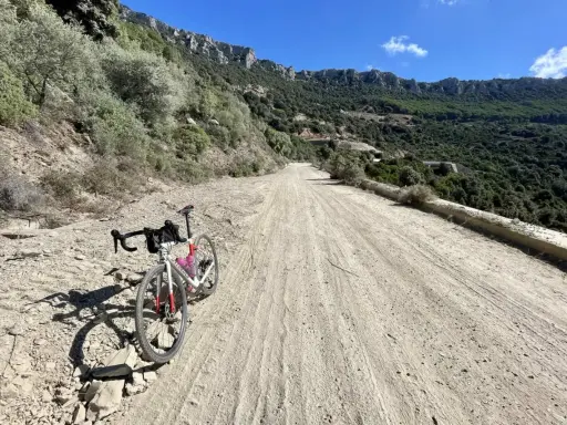 Ein Rennrad steht auf einer staubigen Straße in Sardinien, umgeben von felsigen Hängen und grüner Vegetation.
