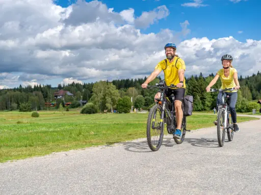 Glückliches Paar auf E-Bikes auf einer Tour durch den Bayerischen Wald. Die Sonne scheint, der Himmel ist blau mit weißen Wolken.