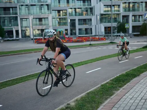 Zwei Rennradfahrerinnen genießen eine Fahrt auf einem Radweg vor modernen Gebäuden.