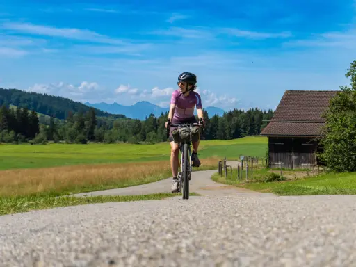 Frau auf einer e bike tour durch grüne Wiesen und Berge, mit einem alten Holzhaus am Wegesrand.