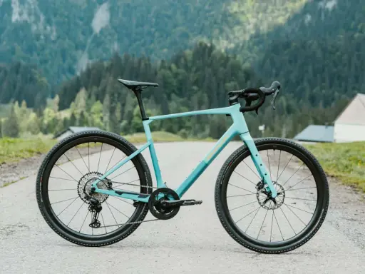 Ein hellblaues Simplon Grid Gravelbike steht auf einer asphaltierten Straße vor einer Bergkulisse mit Wäldern und einer kleinen Kirche.