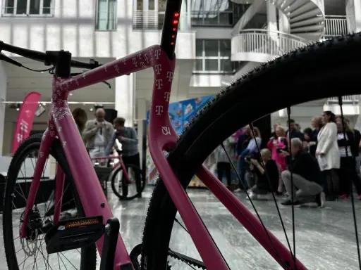 Ein pinkes company bike der Telekom mit vielen T-Logos. Im Hintergrund stehen Menschen und eine Wendeltreppe.