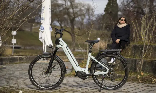 Eine Frau sitzt neben einem hellgrauen Velo de Ville, das auf einem gepflasterten Weg steht. Gute Velo de Ville Erfahrungen sind garantiert.