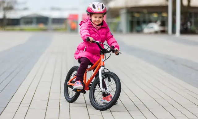 Ein fröhliches Mädchen in pinker Jacke und Helm sitzt auf einem orangefarbenen Kinderfahrrad. Die Frage "kinderfahrrad 4 jahre wieviel zoll" wird durch die Größe des Fahrrads beantwortet.