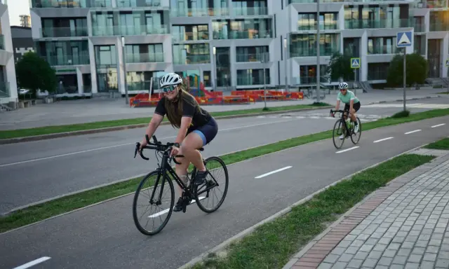 Zwei Rennradfahrerinnen genießen eine Fahrt auf einem Radweg vor modernen Gebäuden.