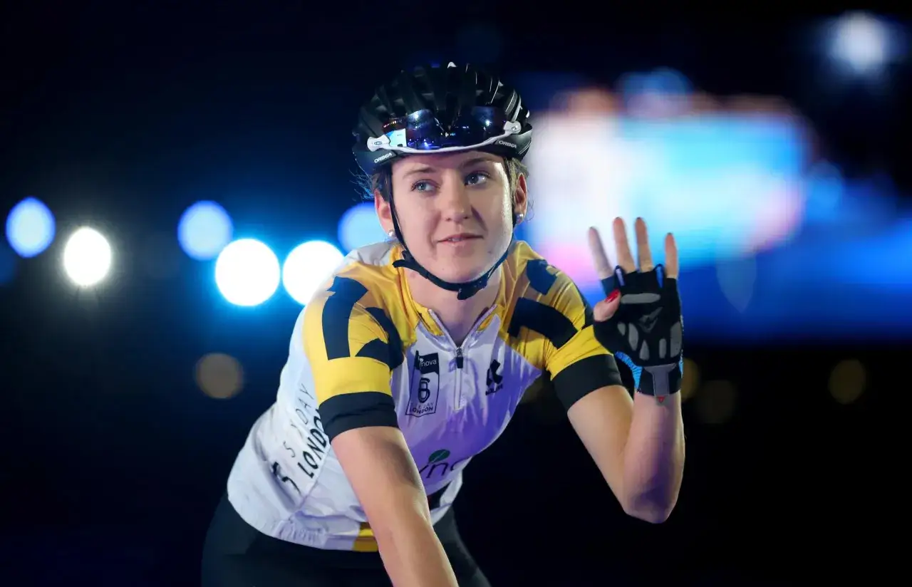 Lea Lin Teutenberg winkt mit der Hand, trägt Radsportkleidung und Helm. Blaue und weiße Lichter im Hintergrund.