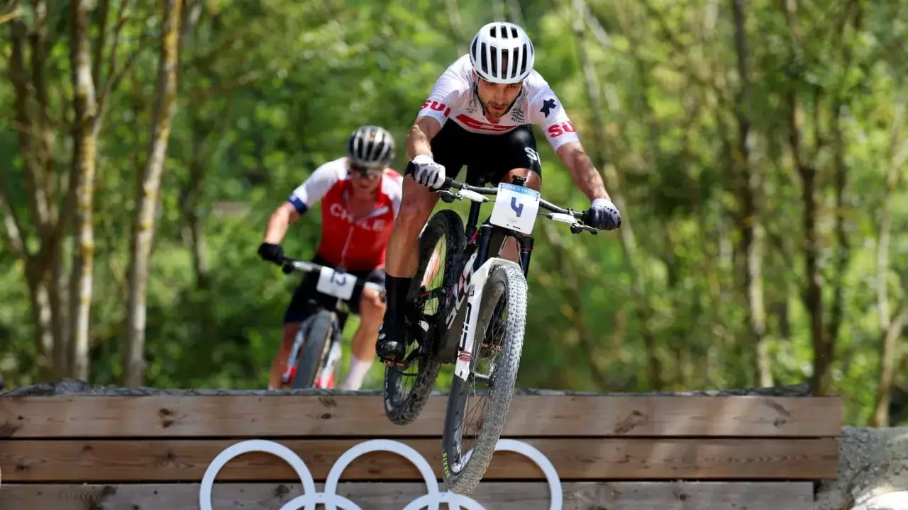 Nino Schurter springt über ein Hindernis beim Mountainbiken. Ein weiterer Fahrer ist im Hintergrund zu sehen.
