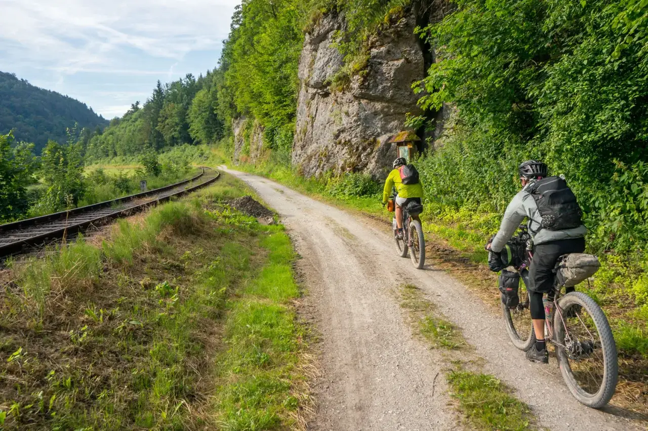 Zwei Radfahrer auf Schotterwegen neben Bahngleisen, umgeben von üppiger Natur.