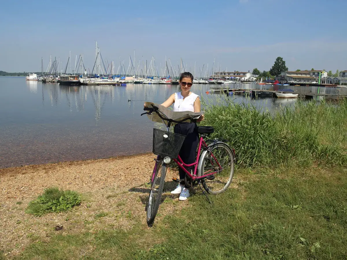 Frau mit Fahrrad am See, plant ihre Radtour Leipzig. Viele Segelboote im Hintergrund.