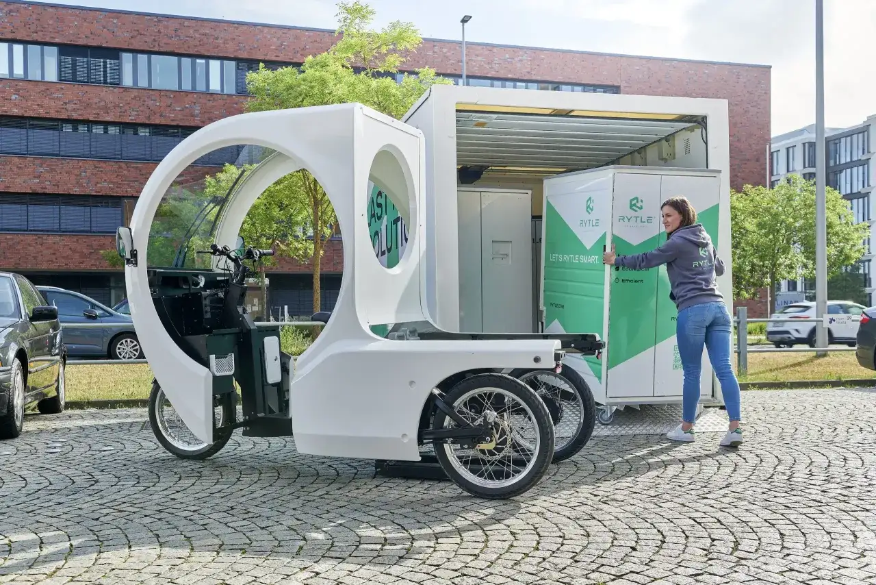 Eine Frau lädt eine Kiste in einen Rytle Movr3. Das Fahrzeug ist dreirädrig und hat eine weiße, futuristische Karosserie.