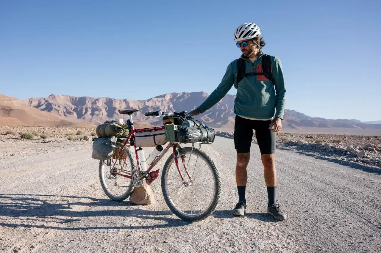 Ein Mann mit Helm und Rucksack steht neben seinem voll beladenen Trekkingrad in einer kargen Berglandschaft.