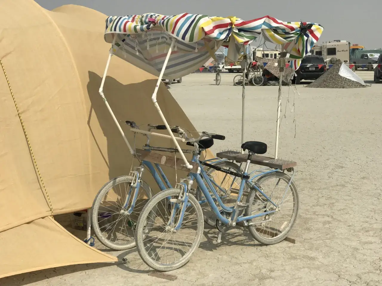 Zwei blaue Fahrräder, ein Buddy Bike, stehen unter einem bunten Sonnenschirm neben einem Zelt.