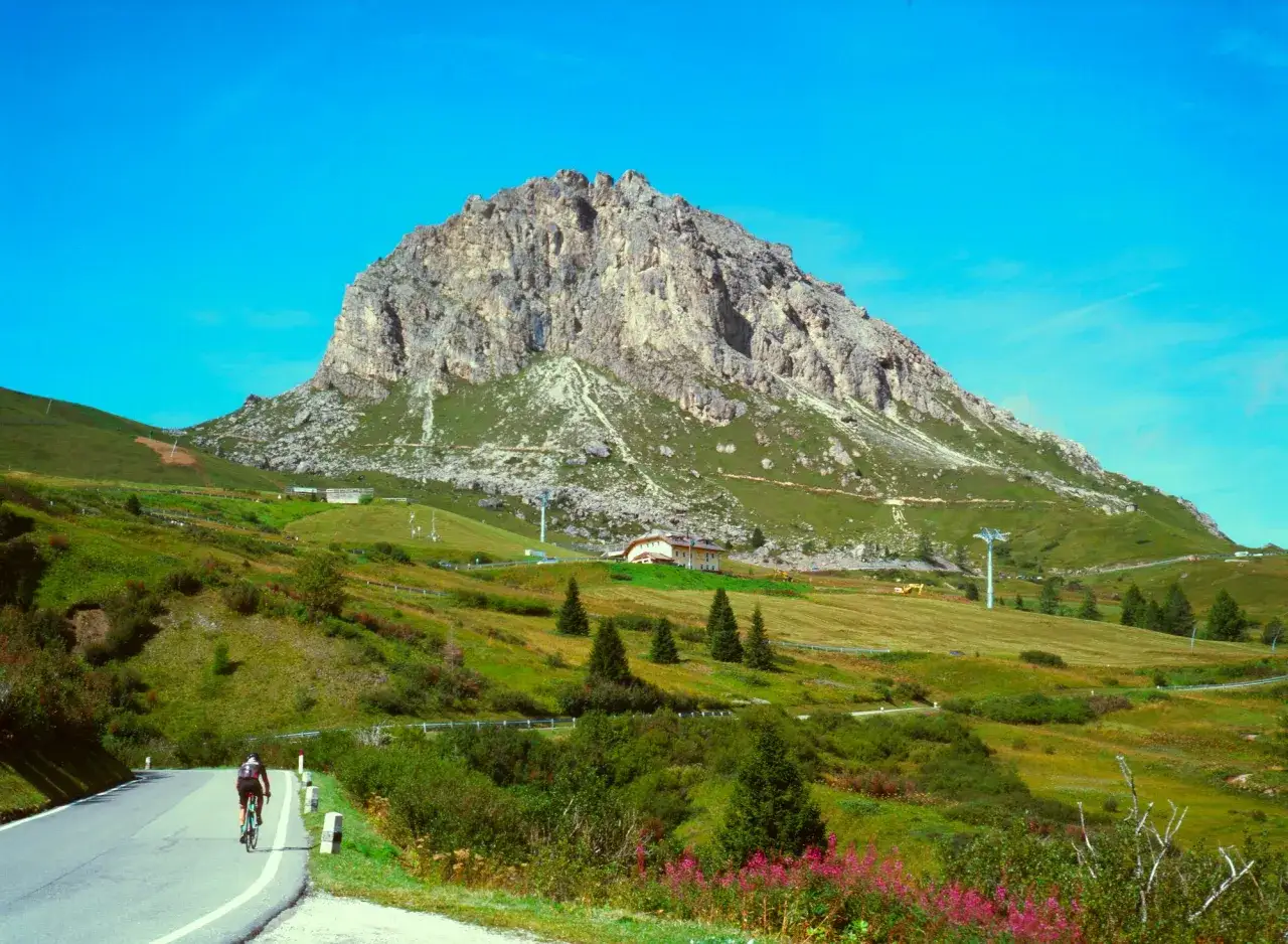 Radfahrer auf einer Bergstraße, die sich dem imposanten Felsmassiv nähert. Die Landschaft ist grün und die Etappe 19 des Giro d'Italia verspricht eine Herausforderung.