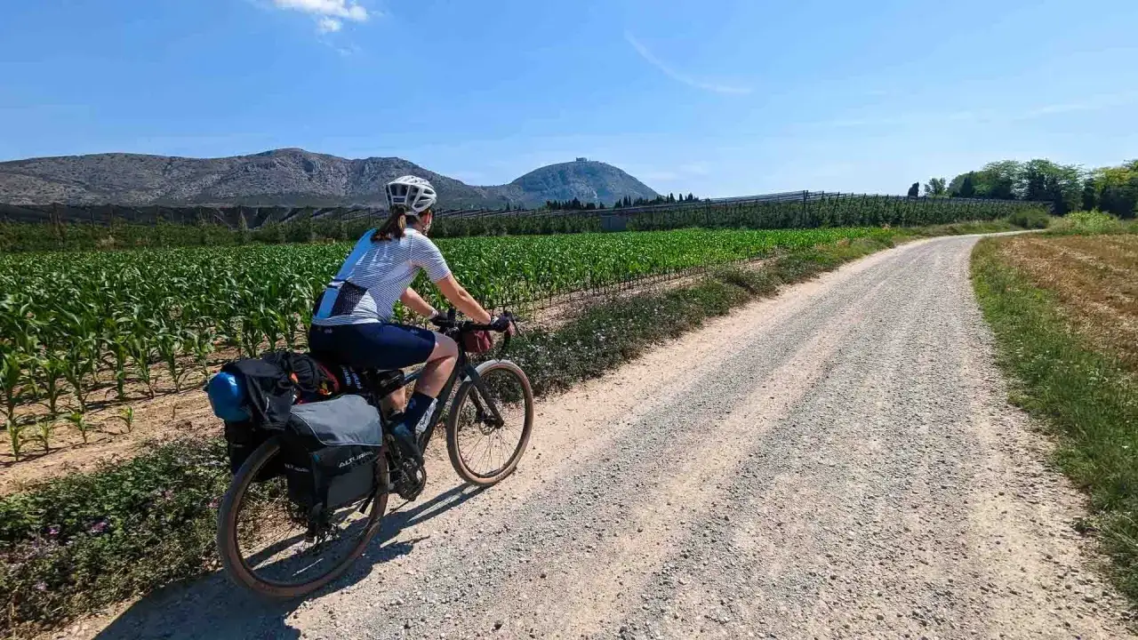 Radfahrerin auf einer Schottertour, vorbei an Maisfeldern und Weinbergen, mit Bergpanorama im Hintergrund.