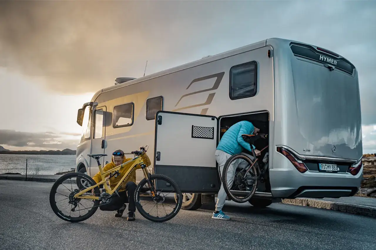 Zwei Personen beladen einen Hymer Wohnwagen mit Fahrr&auml;dern. Ein gelbes Mountainbike steht bereit, w&auml;hrend das andere e bike im Wohnwagen verstaut wird.