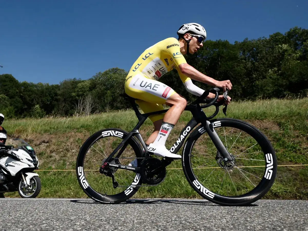 Tadej Pogačar im gelben Trikot fährt auf seinem Rad, die Sonne scheint, im Hintergrund grüne Hügel.