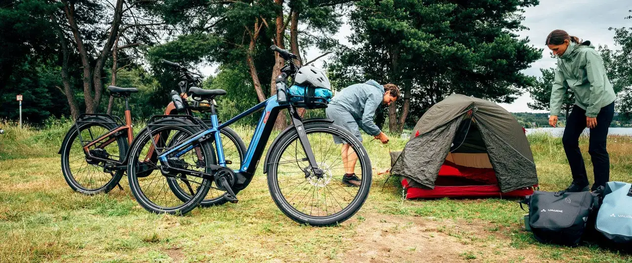 Zwei E-Bikes stehen bereit für eine Tour. Ein Mann baut ein Zelt auf, während eine Frau daneben steht. Campingausrüstung und Taschen sind zu sehen.