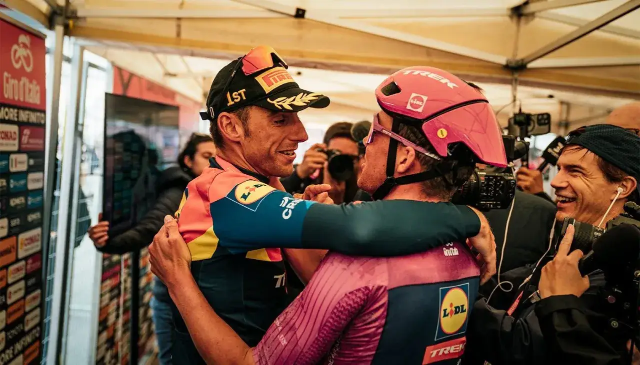 Carlos Verona feiert seinen Sieg beim Giro d'Italia, umarmt von einem Teamkollegen, während Journalisten und Kameras sie umringen.