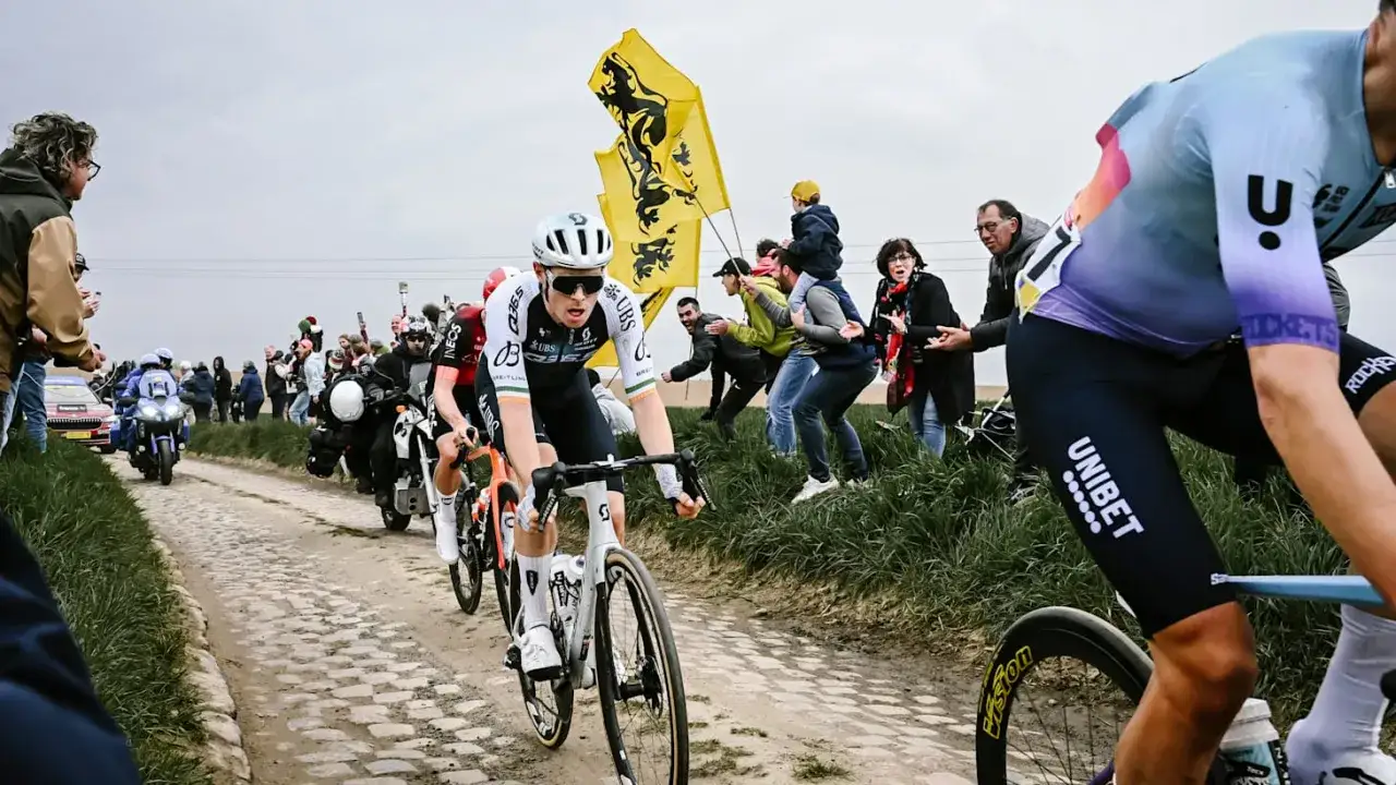 Radrennen Paris-Roubaix: Radfahrer kämpfen sich über Kopfsteinpflaster, während Zuschauer mit Fahnen anfeuern.