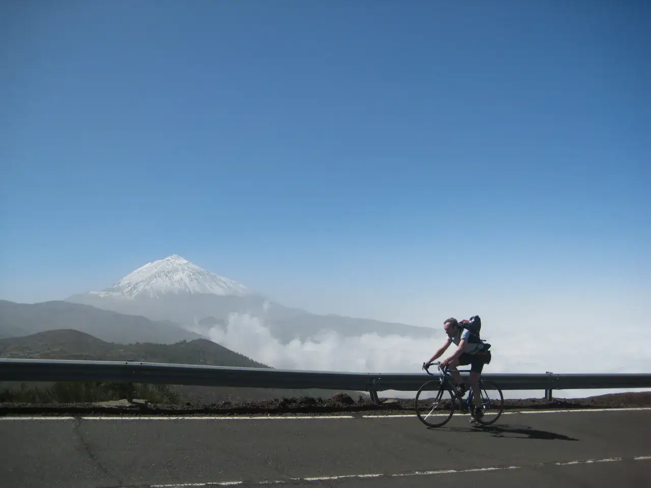 Ein Radfahrer auf Teneriffa mit Rennrad und Rucksack fährt auf einer Straße. Im Hintergrund thront der schneebedeckte Teide.