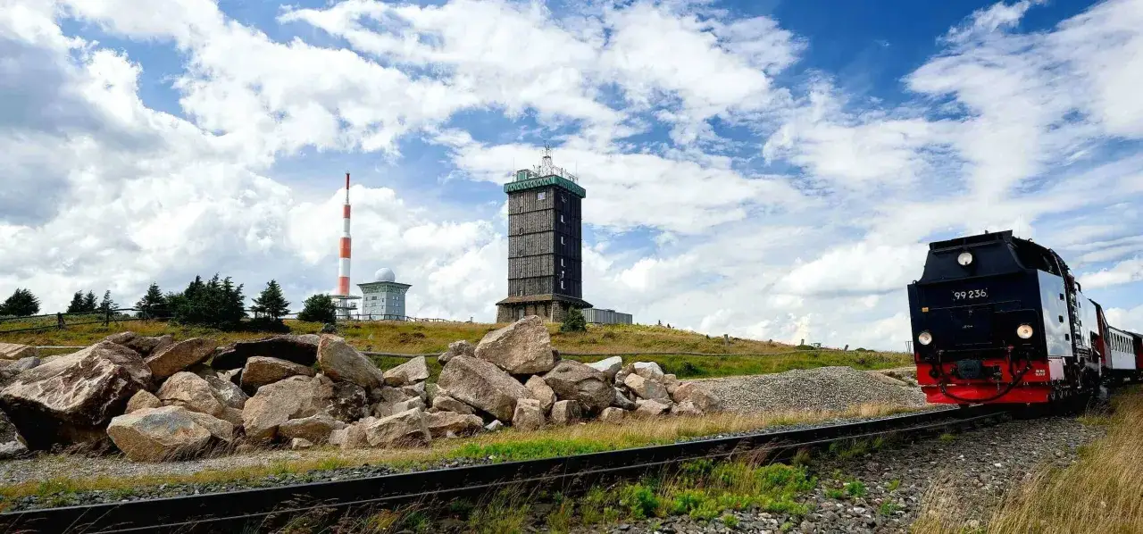 Mit dem e-bike auf den Brocken: Dampflok fährt auf Schienen am Fuße des Turms vorbei, der rote Sendemast im Hintergrund.