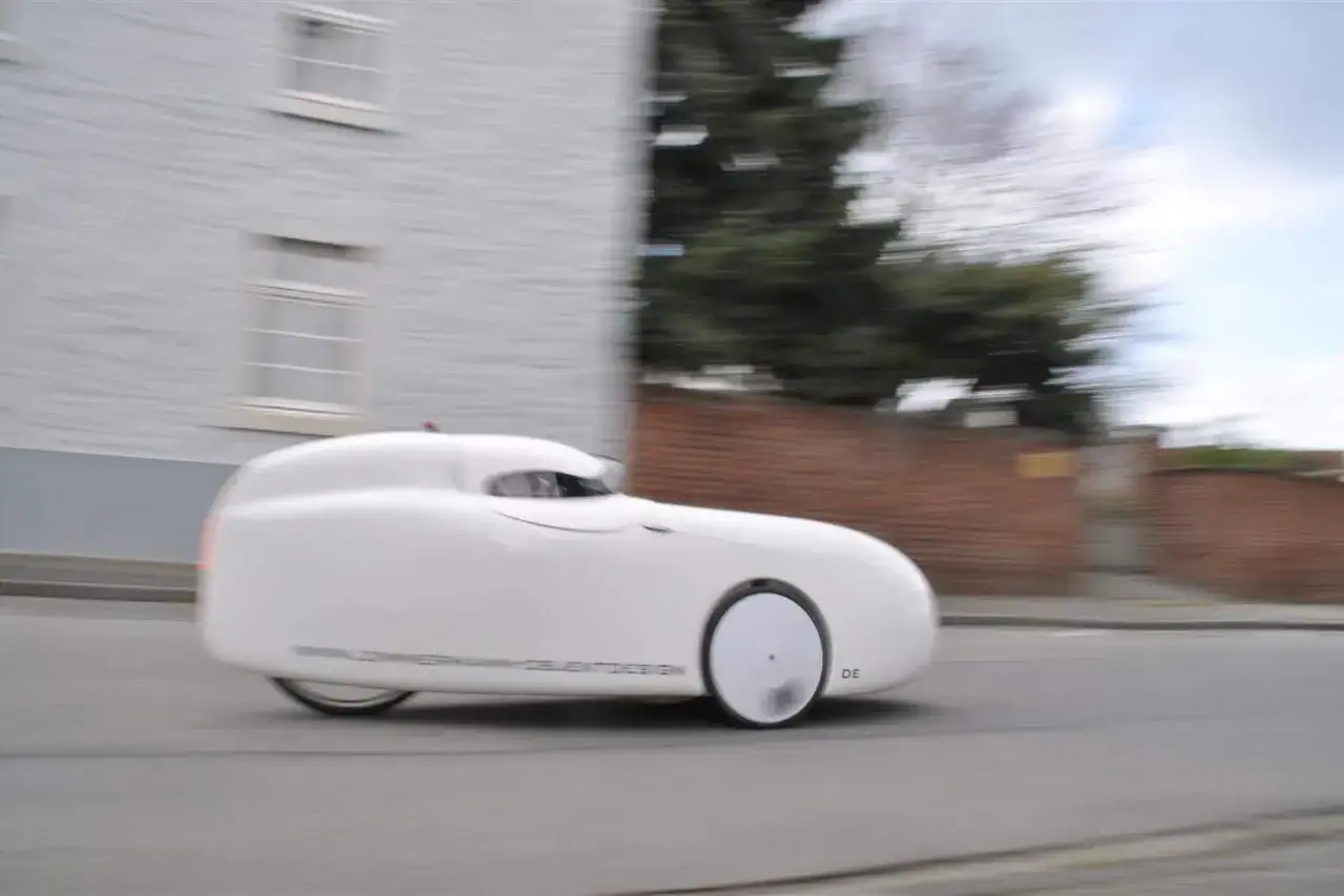 Ein weißer Podbike Frikar fährt auf einer Straße, umgeben von Gebäuden und Bäumen.