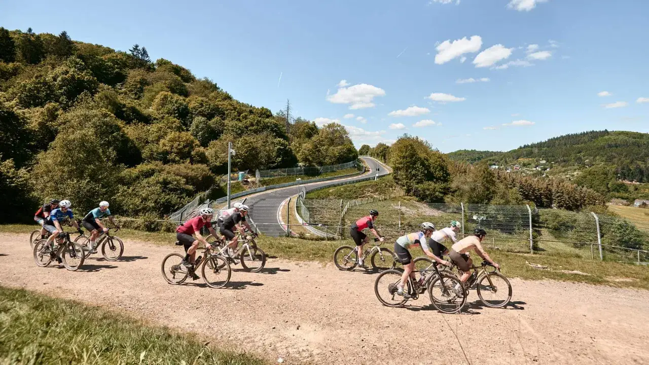 Radfahrer auf einem Schotterweg, der sich durch eine hügelige Landschaft schlängelt. Sie fahren bergauf, die Sonne scheint.