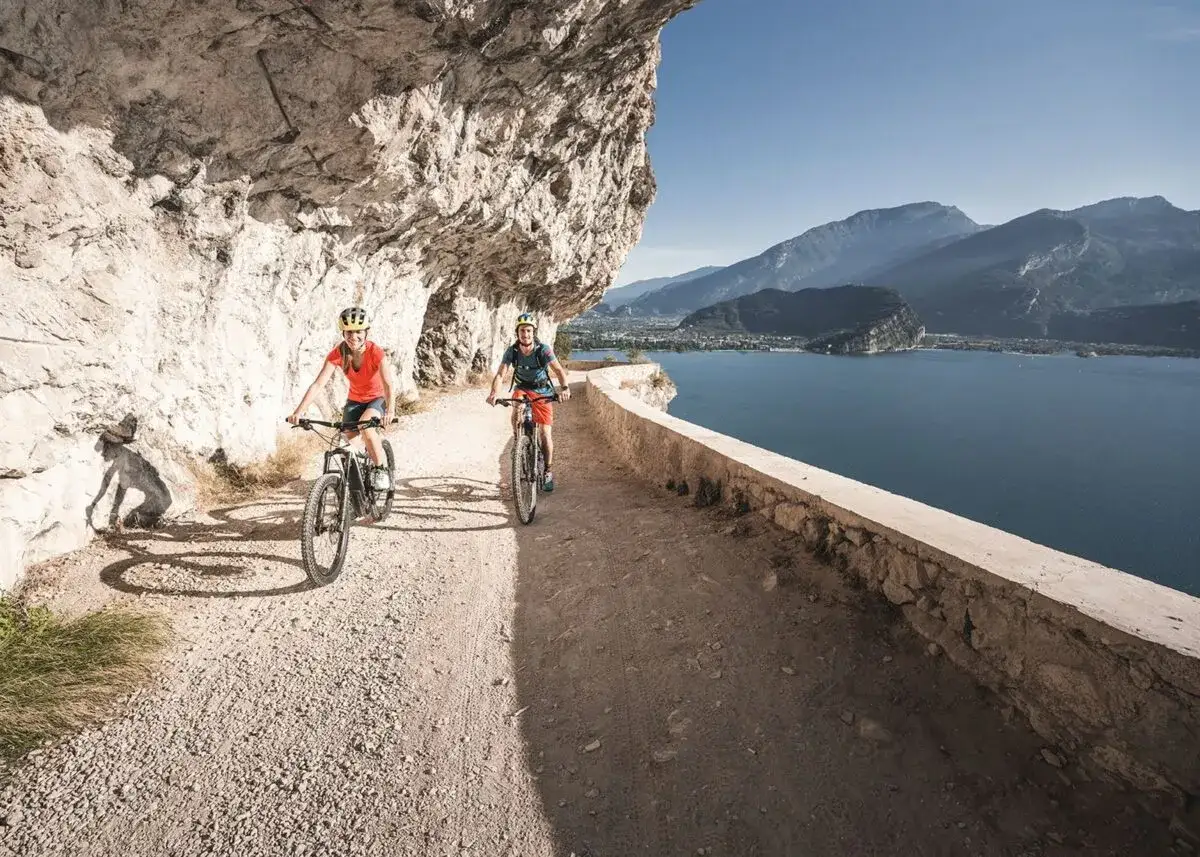 Zwei Radfahrer genießen eine E-Bike Tour am Gardasee auf einem schmalen Weg entlang einer Felswand mit Blick auf den See und die Berge.