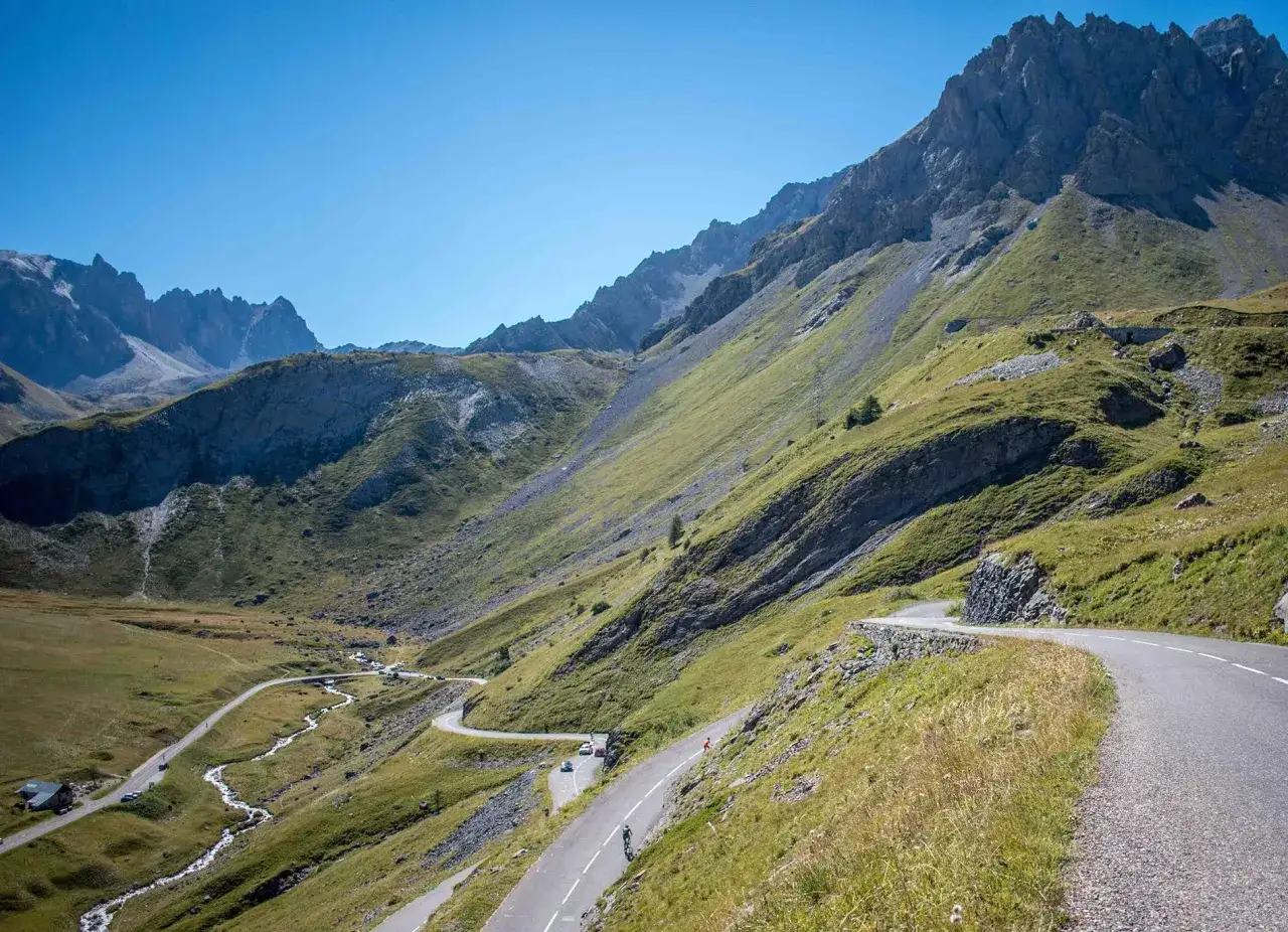 Serpentinenstraße am Col du Galibier schlängelt sich durch grüne Hänge unter majestätischen Bergen. Ein Radfahrer kämpft sich bergauf.