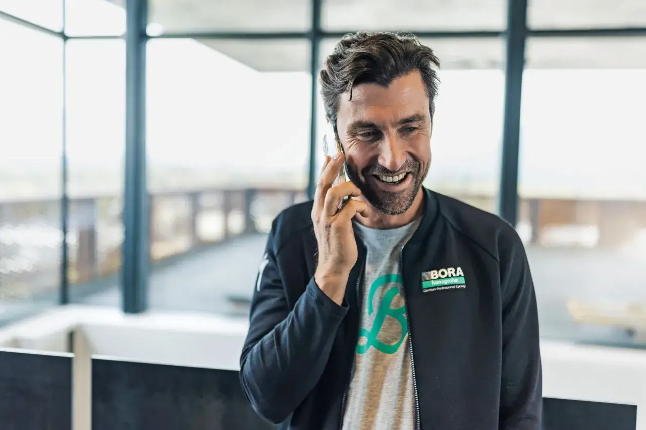 Bernie Eisel lacht am Telefon, trägt eine Jacke mit BORA-hansgrohe Logo.
