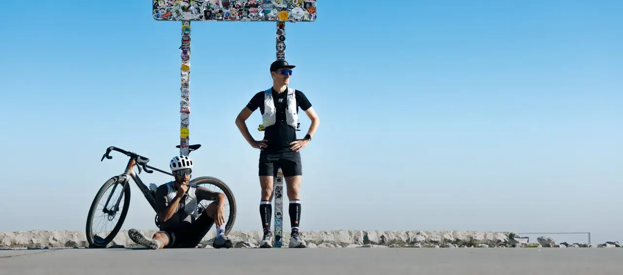 Zwei Radsportler pausieren unter einem mit Stickern beklebten Schild. Einer sitzt neben seinem Rad, der andere steht. Inspiration für die Flandern Rundfahrt.