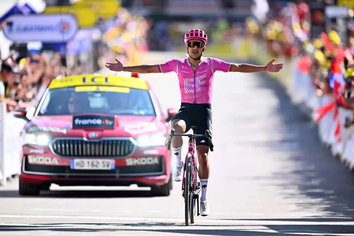 Richard Carapaz feiert seinen Sieg, die Arme weit ausgebreitet, in seinem pinken Trikot. Ein Rennwagen fährt im Hintergrund.