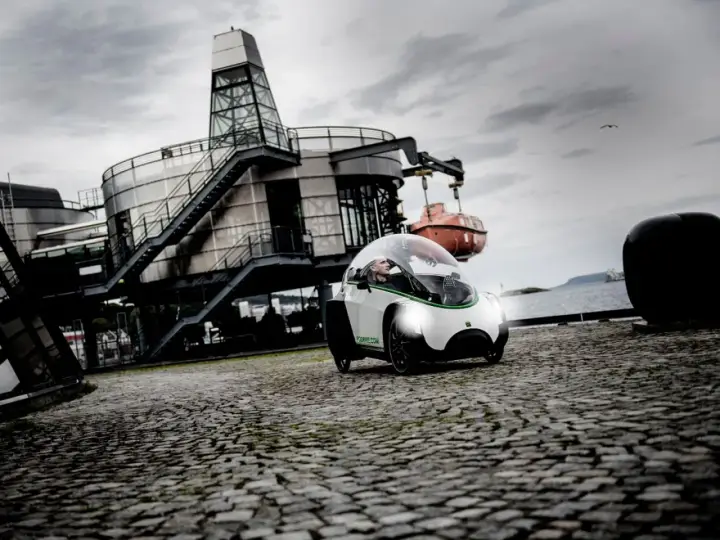 Ein Podbike Frikar fährt auf Kopfsteinpflaster vor einem modernen Gebäude am Wasser.